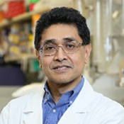 Bangladeshi American Deputy Chief and Senior Investigator, Dr. Javed Khan in his lab, wearing a white lab coat, standing with his arms folded in front of him.