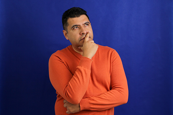 A curious Hispanic/Latino man stares with his hand over his mouth stands in front of a blue background. 