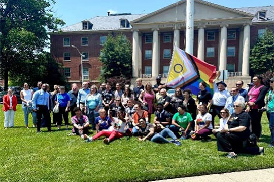 A group photo of Salutaris members in front of NIH Building 1 with the Progress Pride flag behind the group. 