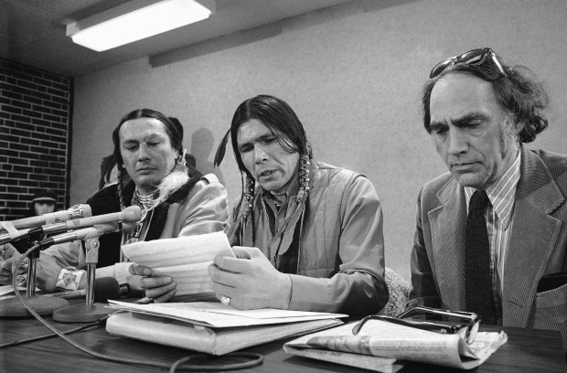 Three people sit at a table with microphones on it reading a statement from a piece of paper.