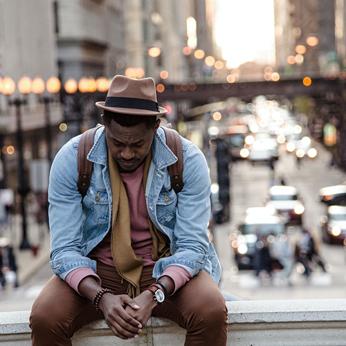 A Black man with his head held down sitting on an overpass, with a city landscape in the background