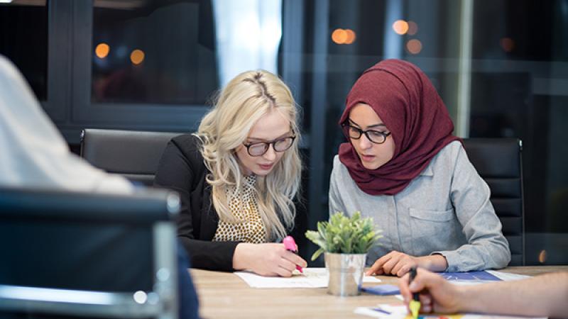 A blonde woman wearing glasses working beside another woman wearing glasses and a headscarf at an office desk.