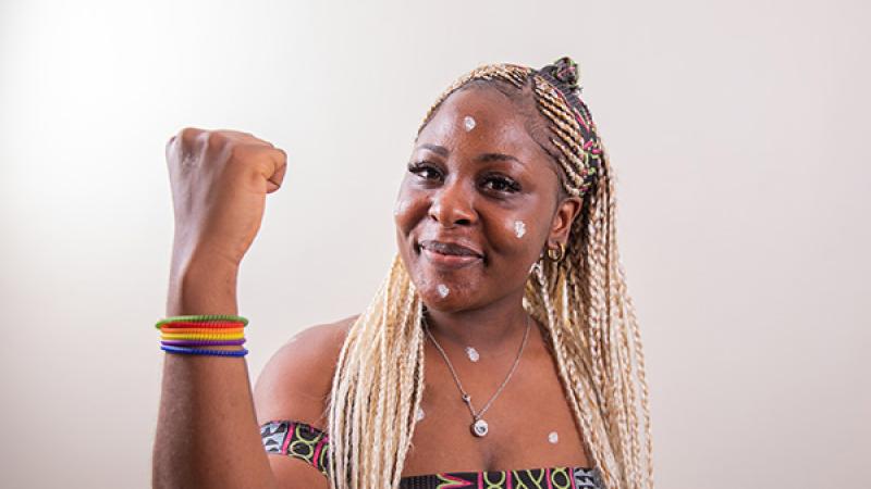 A Black lesbian woman with her arm raised shows her bicep and wears colorful, rainbow colored bracelets. She wears an African print halter top with black, green, purple, and pink geometric shapes.