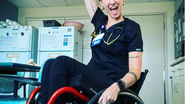 Ryann Mason in black scrubs sits in a wheelchair and pumps fist with an excited expression on her face.